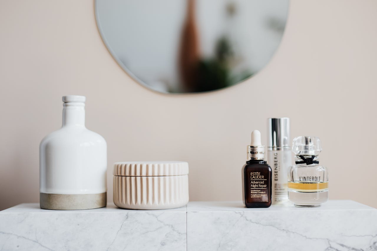 Glass flasks with cream and lotion near perfume and big ceramic vase on marble surface close to beige wall with oval shaped mirror